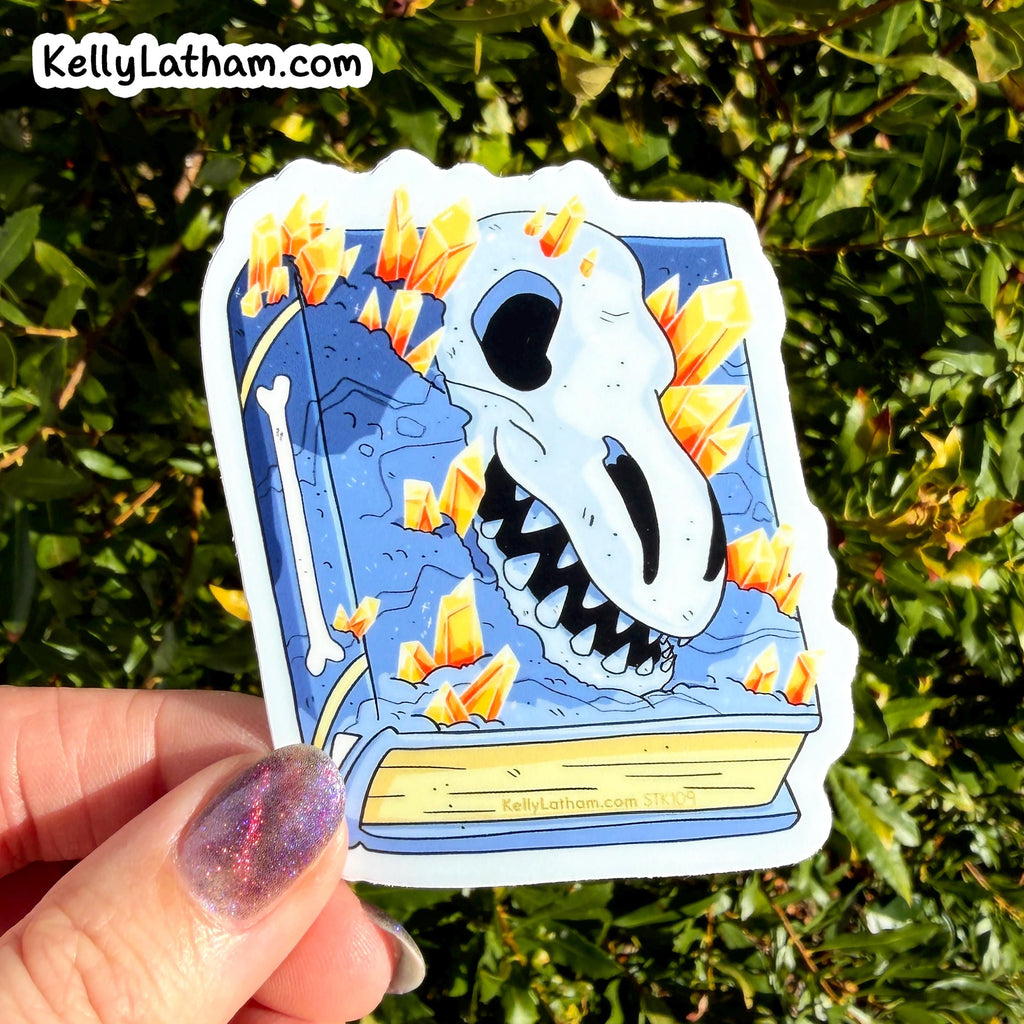 A hand with glittery purple nail polish holds a die-cut sticker featuring a blue book with a white skull and orange crystals, set against a background of green foliage. The sticker has the brand name KellyLatham.