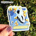 A hand with glittery purple nail polish holds a die-cut sticker featuring a blue book with a white skull and orange crystals, set against a background of green foliage. The sticker has the brand name KellyLatham.