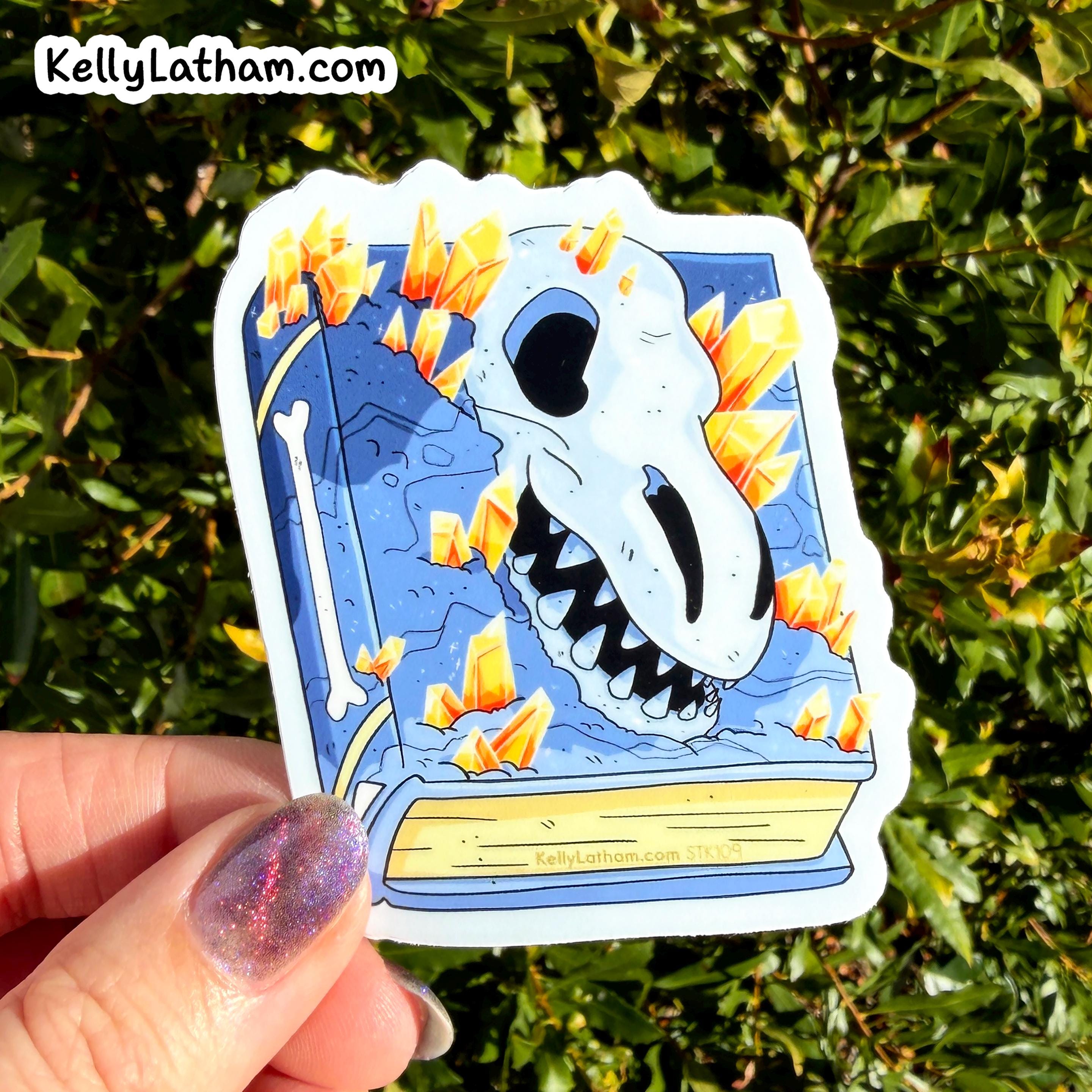 A hand with glittery purple nail polish holds a die-cut sticker featuring a blue book with a white skull and orange crystals, set against a background of green foliage. The sticker has the brand name KellyLatham.