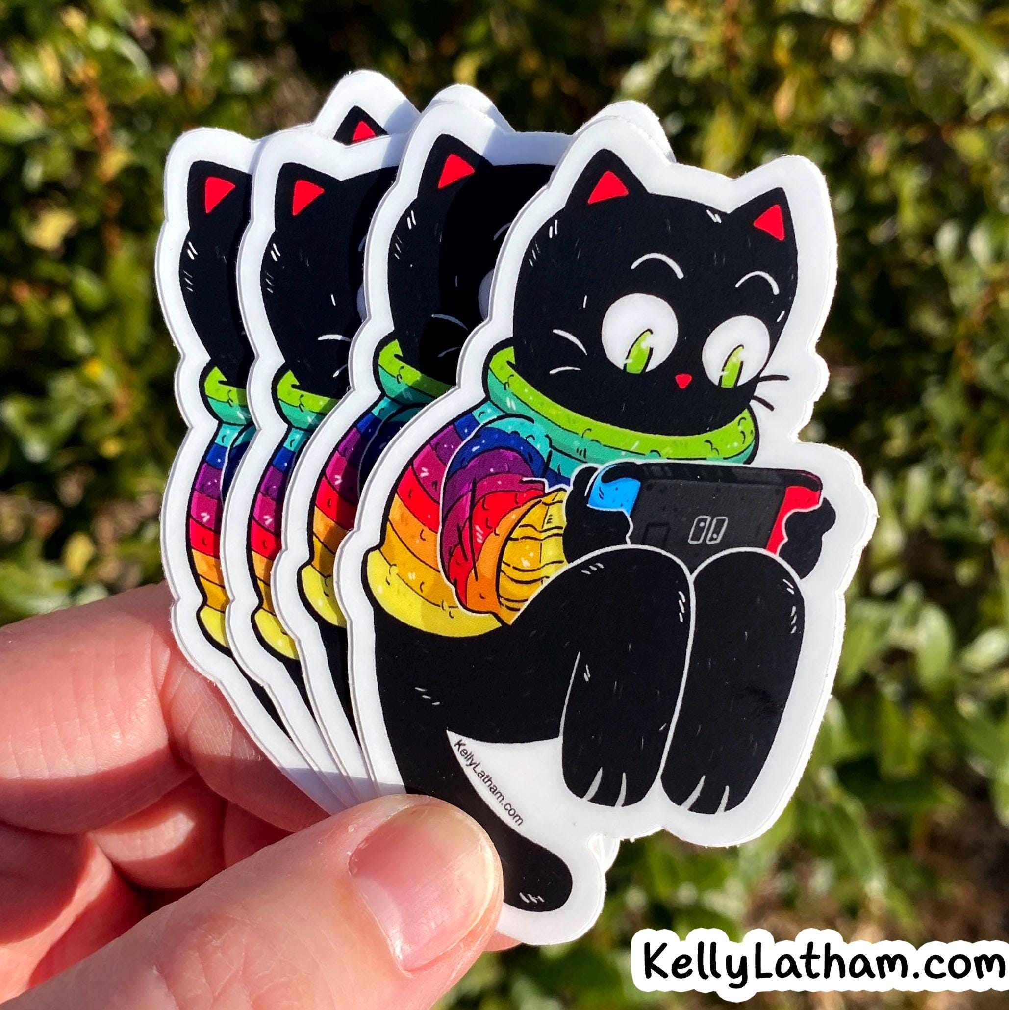 A hand holds a stack of die-cut stickers featuring a black cat wearing a bright rainbow scarf and holding a game controller, photographed outdoors against blurred green foliage. A small 'KellyLatham.