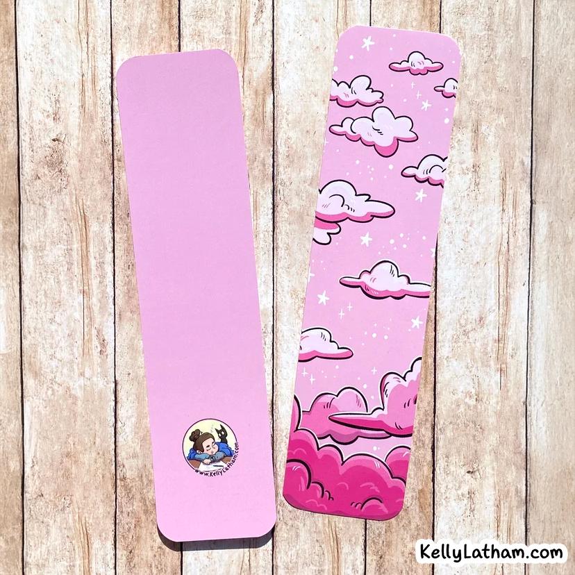 Two pink bookmarks with cloud and star designs are displayed on a light wood background. One bookmark is plain pink with a small circular cartoon logo, and the other has a whimsical pink cloud pattern.