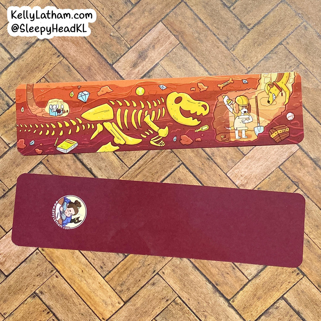 Two rectangular yoga mats laid flat on a wooden herringbone floor. The top mat has a colorful cartoon illustration featuring a yellow dinosaur skeleton and prehistoric-themed artwork on an orange-brown background.