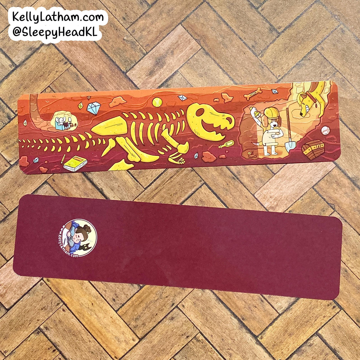 Two rectangular yoga mats laid flat on a wooden herringbone floor. The top mat has a colorful cartoon illustration featuring a yellow dinosaur skeleton and prehistoric-themed artwork on an orange-brown background.