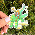 Hand holding a sticker of a person riding a green dinosaur with a person in an orange life jacket against a green leafy background.
