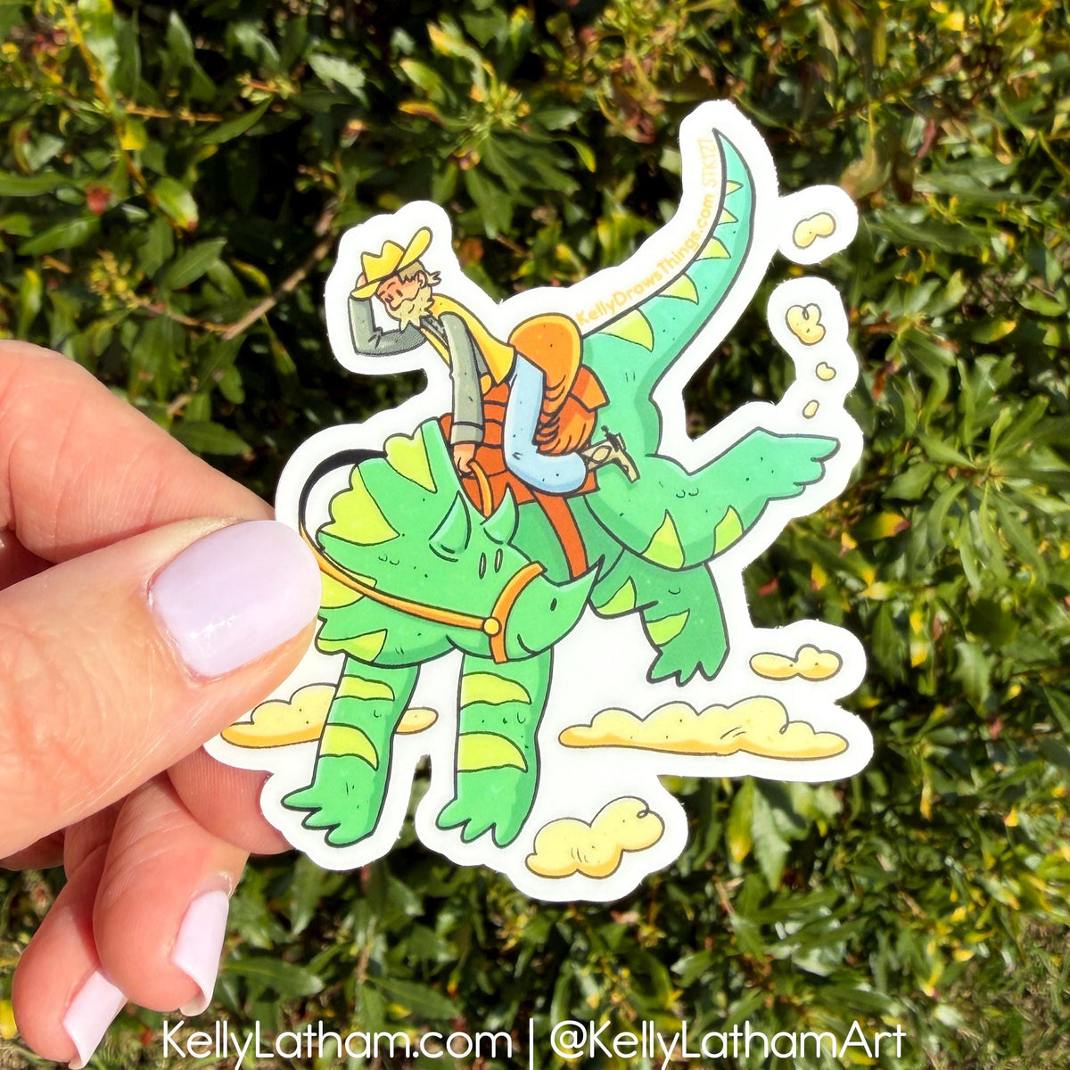 Hand holding a sticker of a person riding a green dinosaur with a person in an orange life jacket against a green leafy background.