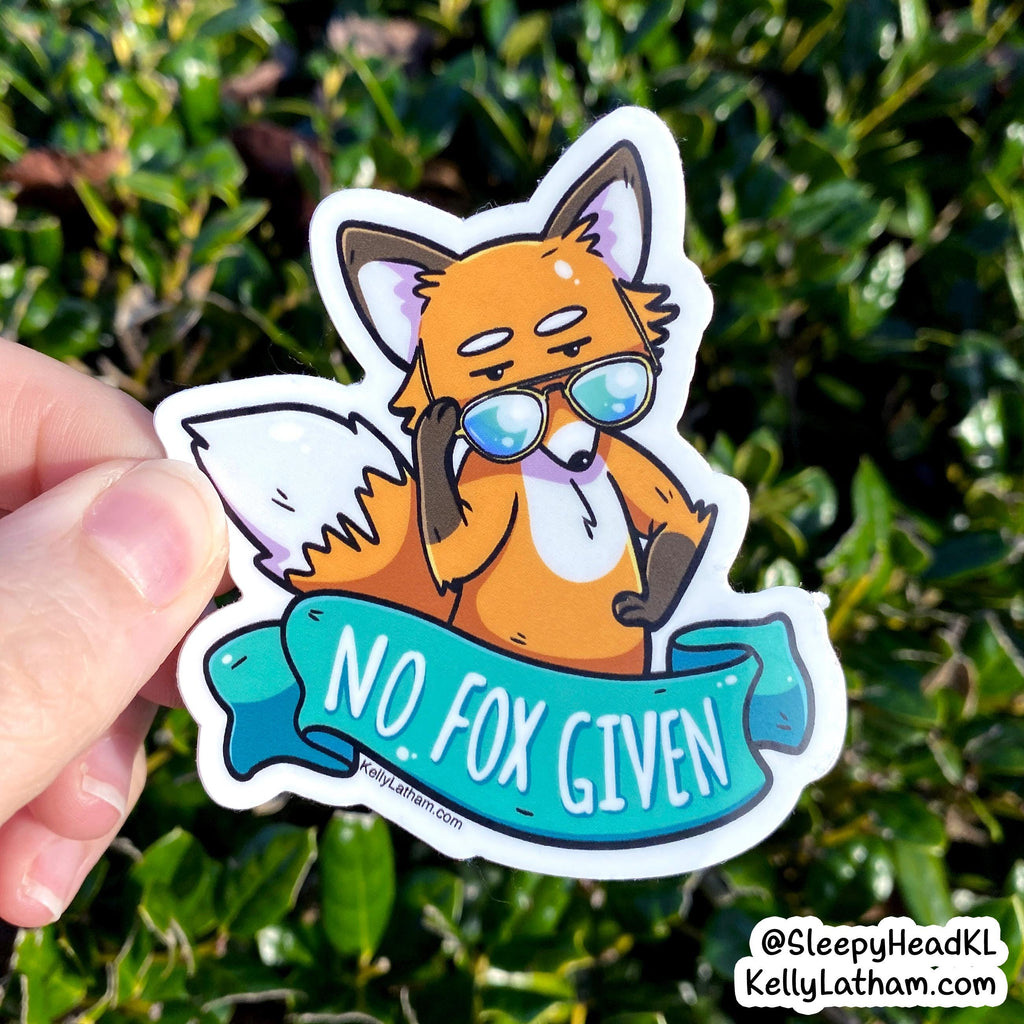A hand holds a die-cut sticker featuring a cartoon orange fox wearing blue-tinted sunglasses with a teal banner that reads “NO. FOX GIVEN.