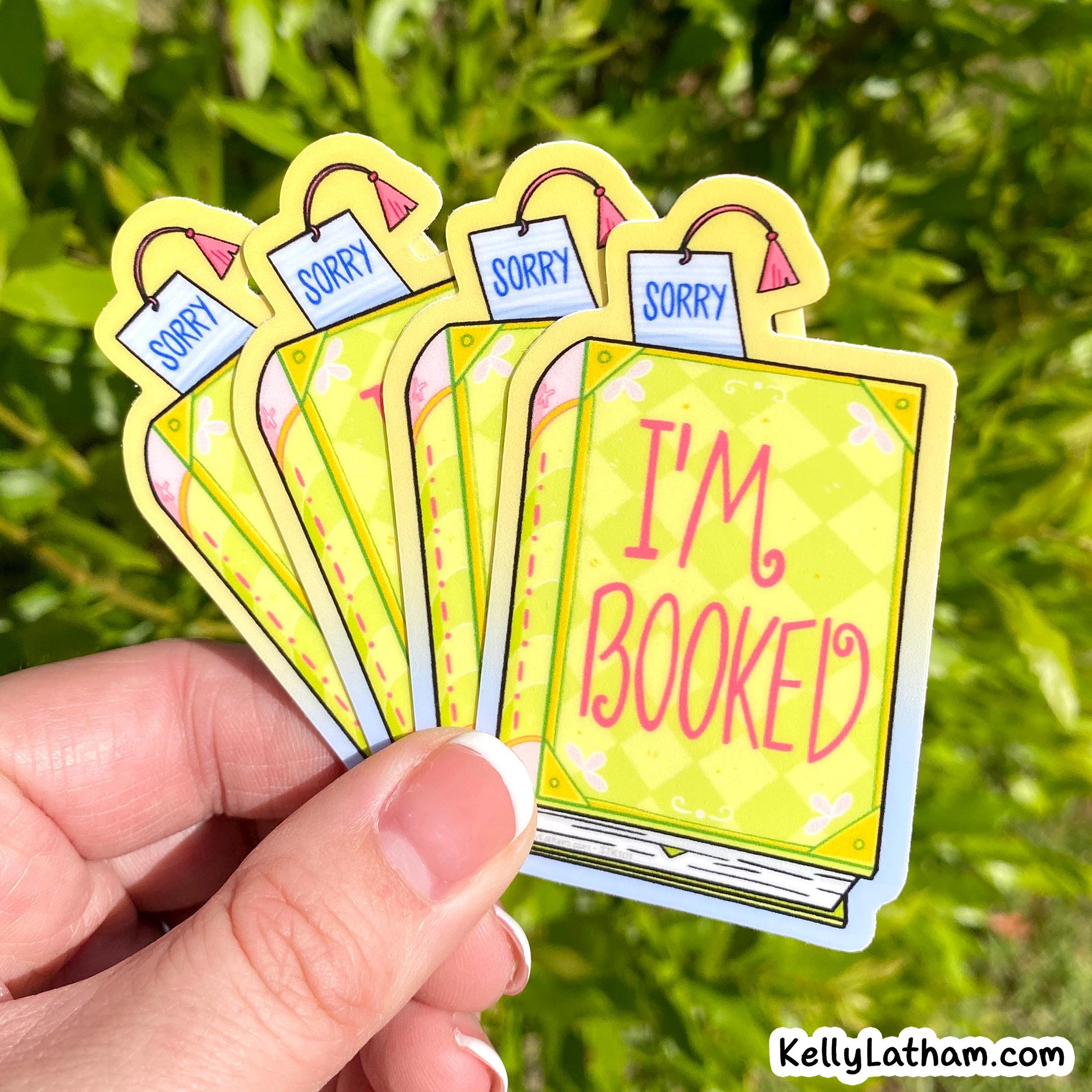 A hand holds a fan of five die-cut stickers shaped like books with price tags, featuring the words 'SORRY' and 'I'M BOOKED' against a green outdoor background.