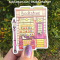 A hand holds a die-cut sticker featuring a whimsical illustrated bookstore with a pink door, yellow storefront, and a pink streetlamp, set against a blurred green outdoor background.