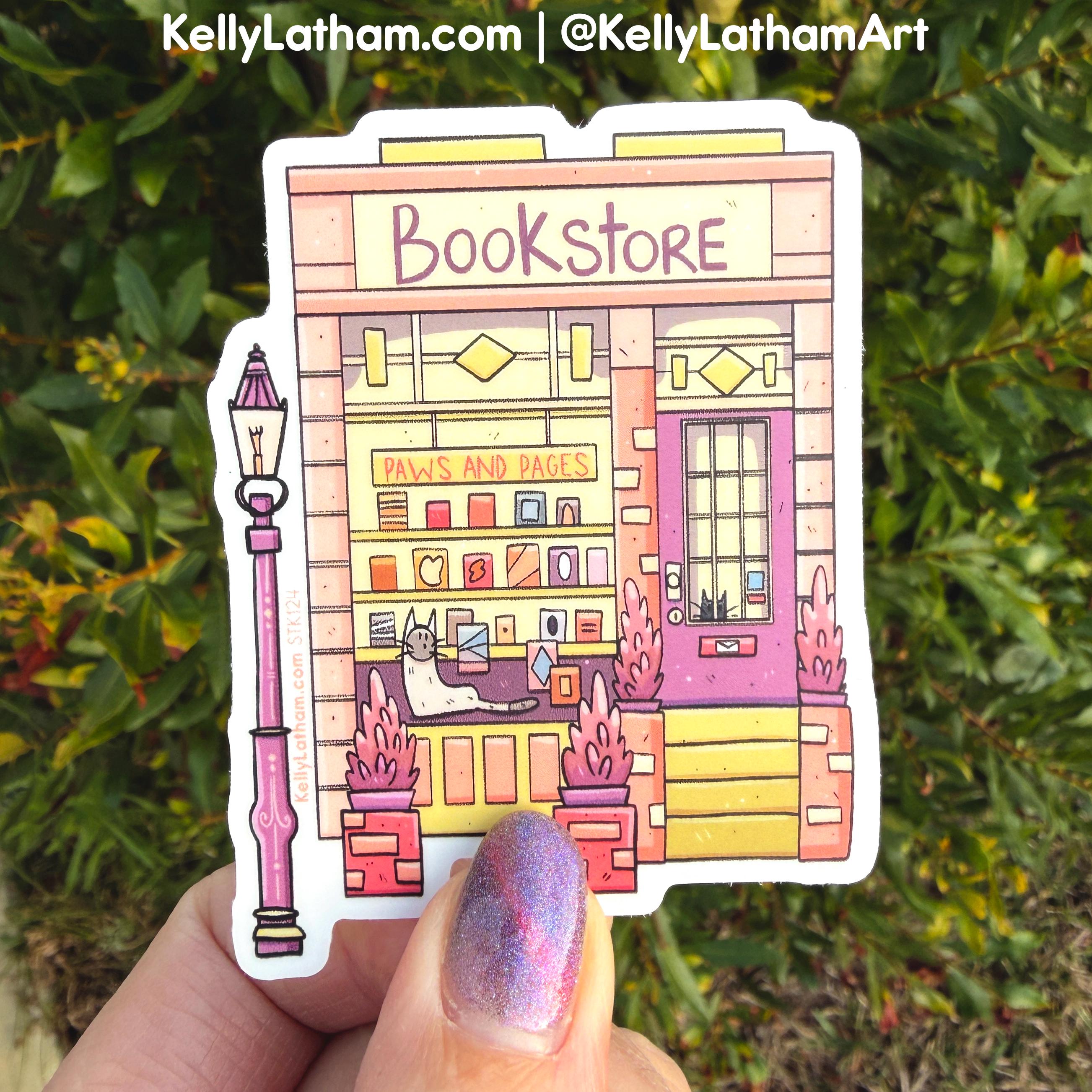 A hand holds a die-cut sticker featuring a whimsical illustrated bookstore with a pink door, yellow storefront, and a pink streetlamp, set against a blurred green outdoor background.