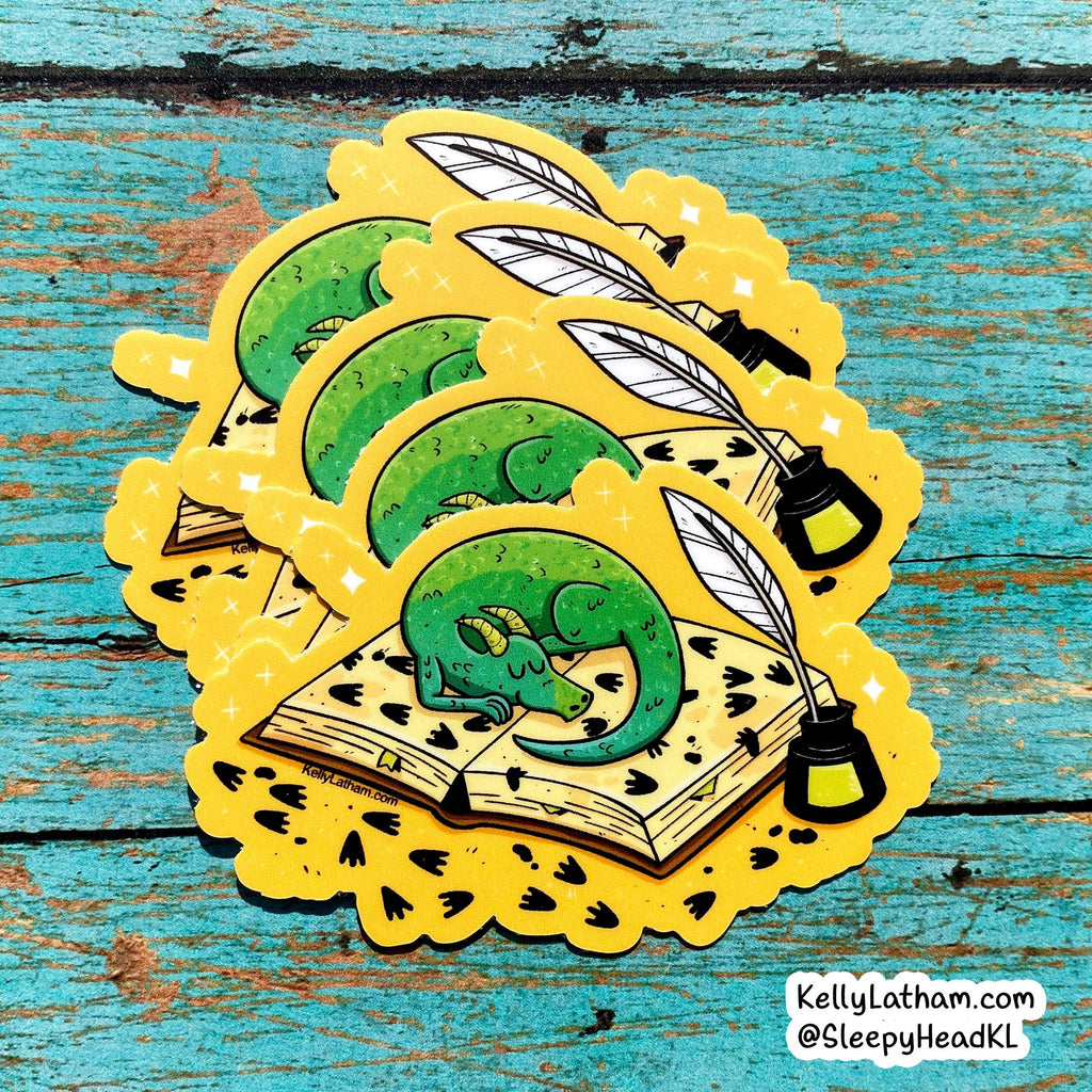 Three die-cut stickers featuring a green sleeping dragon curled up in an open book with a quill pen and ink bottle, arranged on a distressed turquoise wooden surface. The stickers have a bright yellow border with small black heart details.