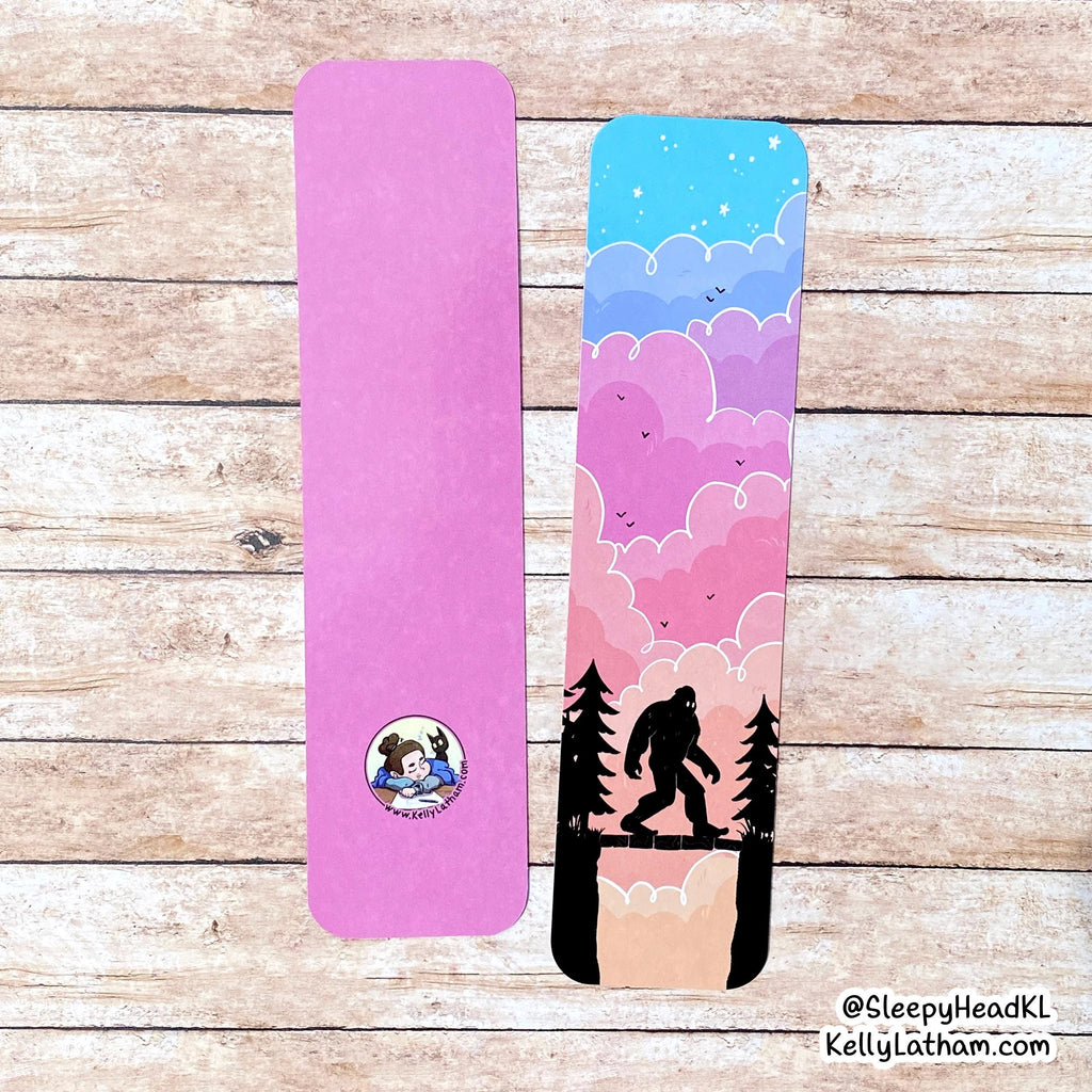 Two rectangular bookmarks of bigfoot silhouette at sunset displayed on a rustic wood plank background. The left piece is solid pink with a small circular cartoon logo near the bottom. 
