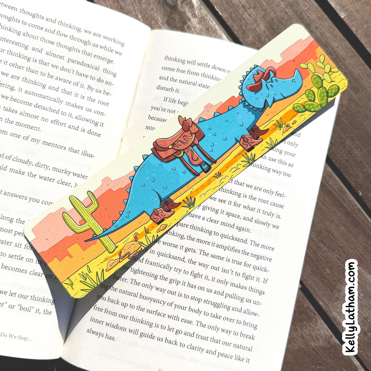 A colorful illustrated bookmark shaped like a desert landscape with a blue dinosaur and a red truck, placed across an open book on a dark wooden surface.