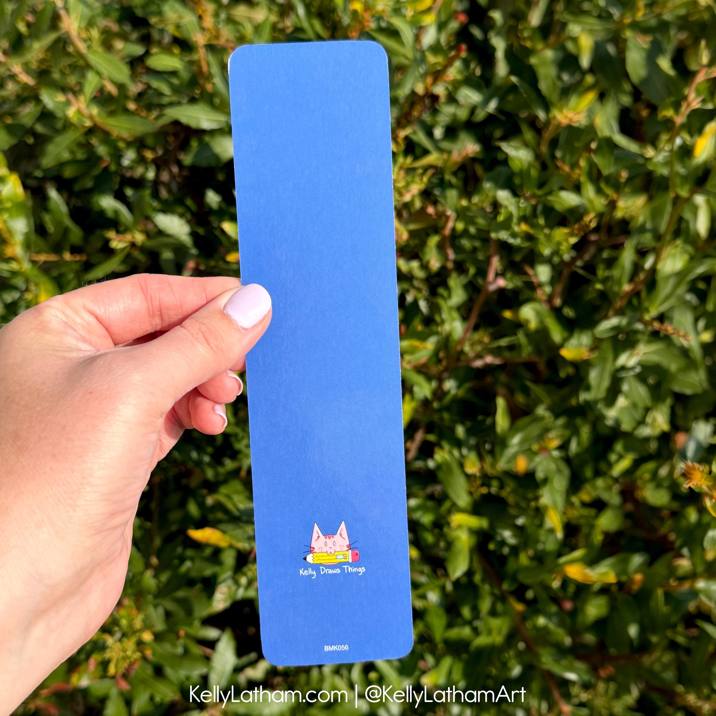 A hand holds a tall, narrow blue art print or bookmark featuring a small pink cat illustration and the text 'Big Dreams Things' against a background of green foliage.