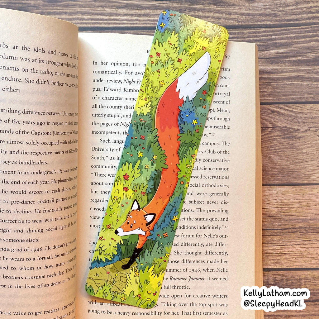 A decorative illustrated book bookmark shaped like a fox sitting on a grassy hill, placed on the pages of an open book on a wooden surface.