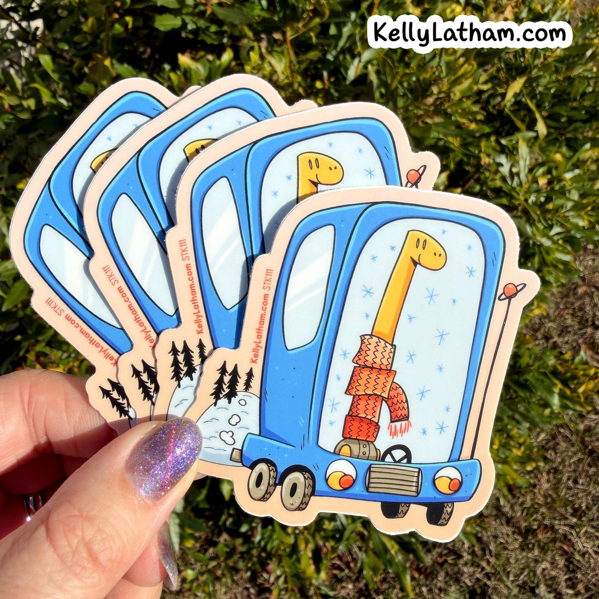 A hand with glittery purple nails holds a fan of four die-cut stickers outdoors against a background of green foliage. The stickers feature a blue truck with a giraffe wearing an orange knit sweater and scarf, with the text 'KellyLatham.