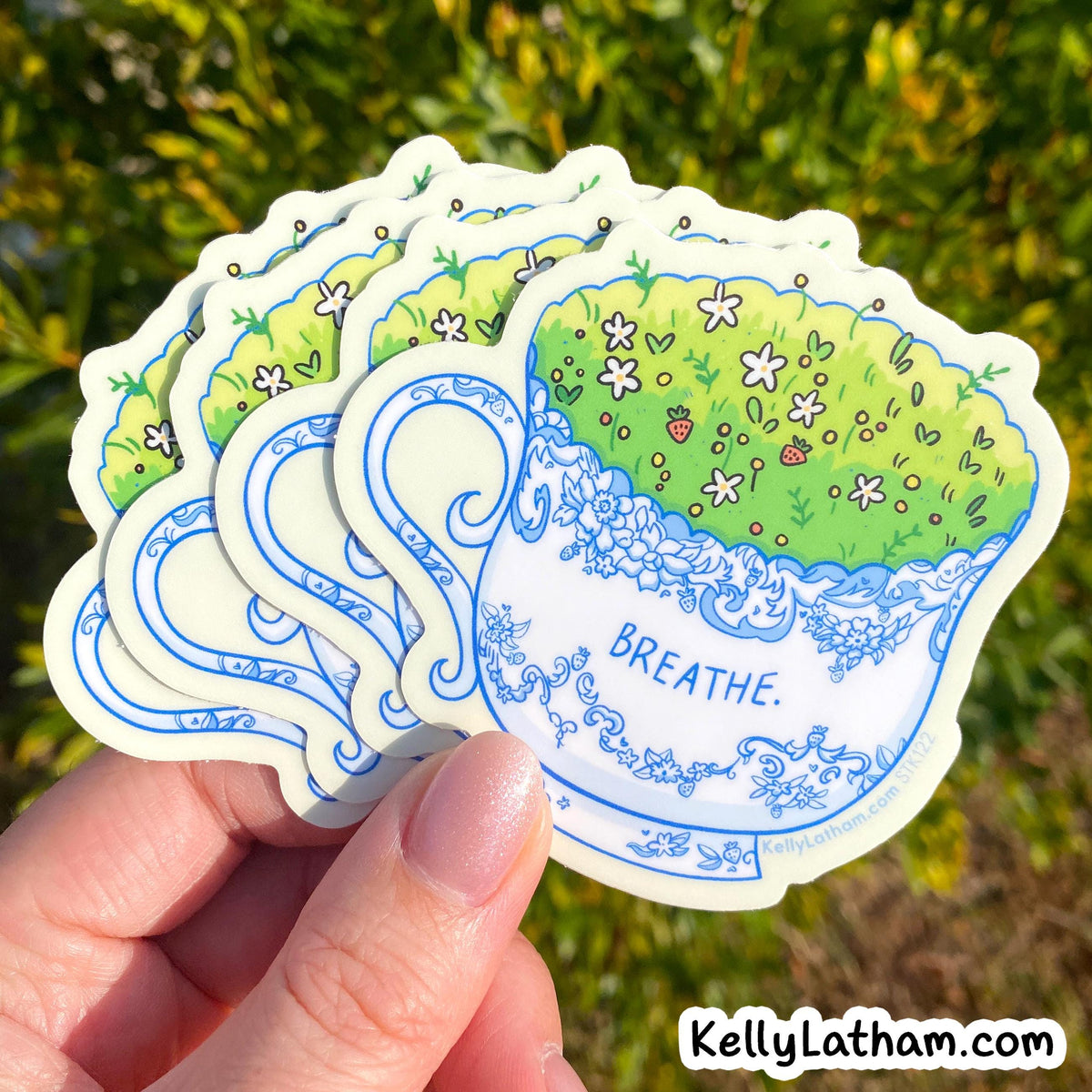 A hand holds a fan of die-cut stickers featuring an illustrated teacup filled with green grass and small white flowers, with the word “BREATHE.” printed on the front.