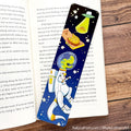 A decorative paper bookmark featuring a cartoon astronaut and a slice of pizza on a black space background, placed on an open book over a wooden surface.