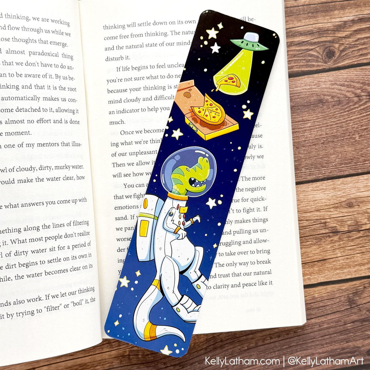 A decorative paper bookmark featuring a cartoon astronaut and a slice of pizza on a black space background, placed on an open book over a wooden surface.