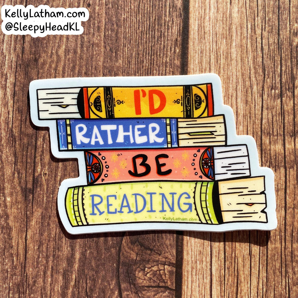 A die-cut sticker shaped like stacked books with the phrase “I’D RATHER BE READING” in colorful lettering, placed on a brown wood surface. The sticker includes a white border and a small visible brand watermark in the top-left corner reading KellyLatham.