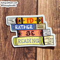 A die-cut sticker shaped like stacked books with the phrase “I’D RATHER BE READING” in colorful lettering, placed on a brown wood surface. The sticker includes a white border and a small visible brand watermark in the top-left corner reading KellyLatham.