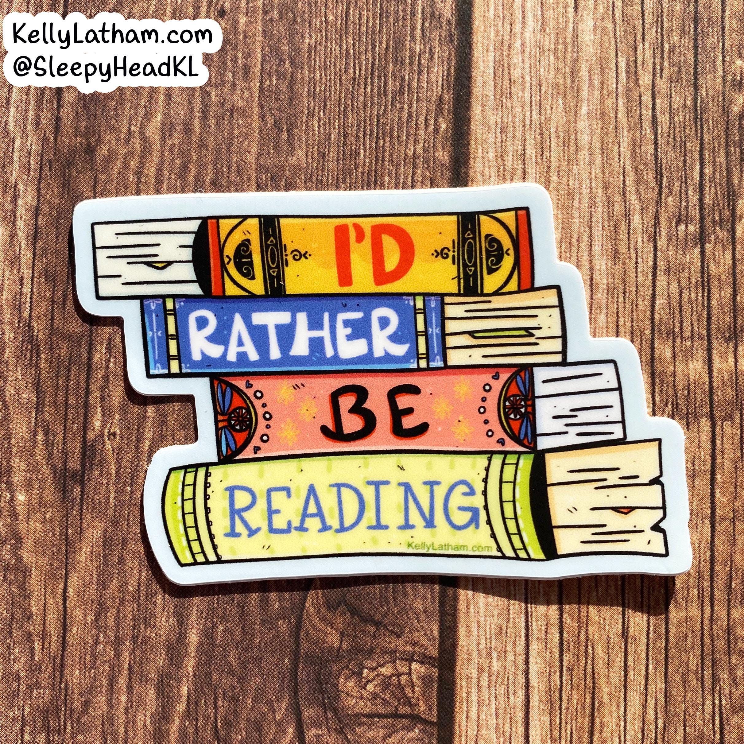A die-cut sticker shaped like stacked books with the phrase “I’D RATHER BE READING” in colorful lettering, placed on a brown wood surface. The sticker includes a white border and a small visible brand watermark in the top-left corner reading KellyLatham.