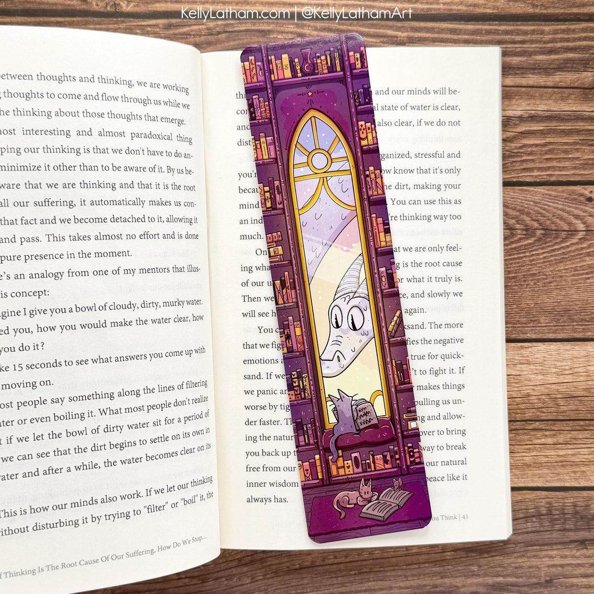 A purple and gold illustrated bookmark or book divider with a cartoon character and book illustrations, placed on an open book on a wooden surface.