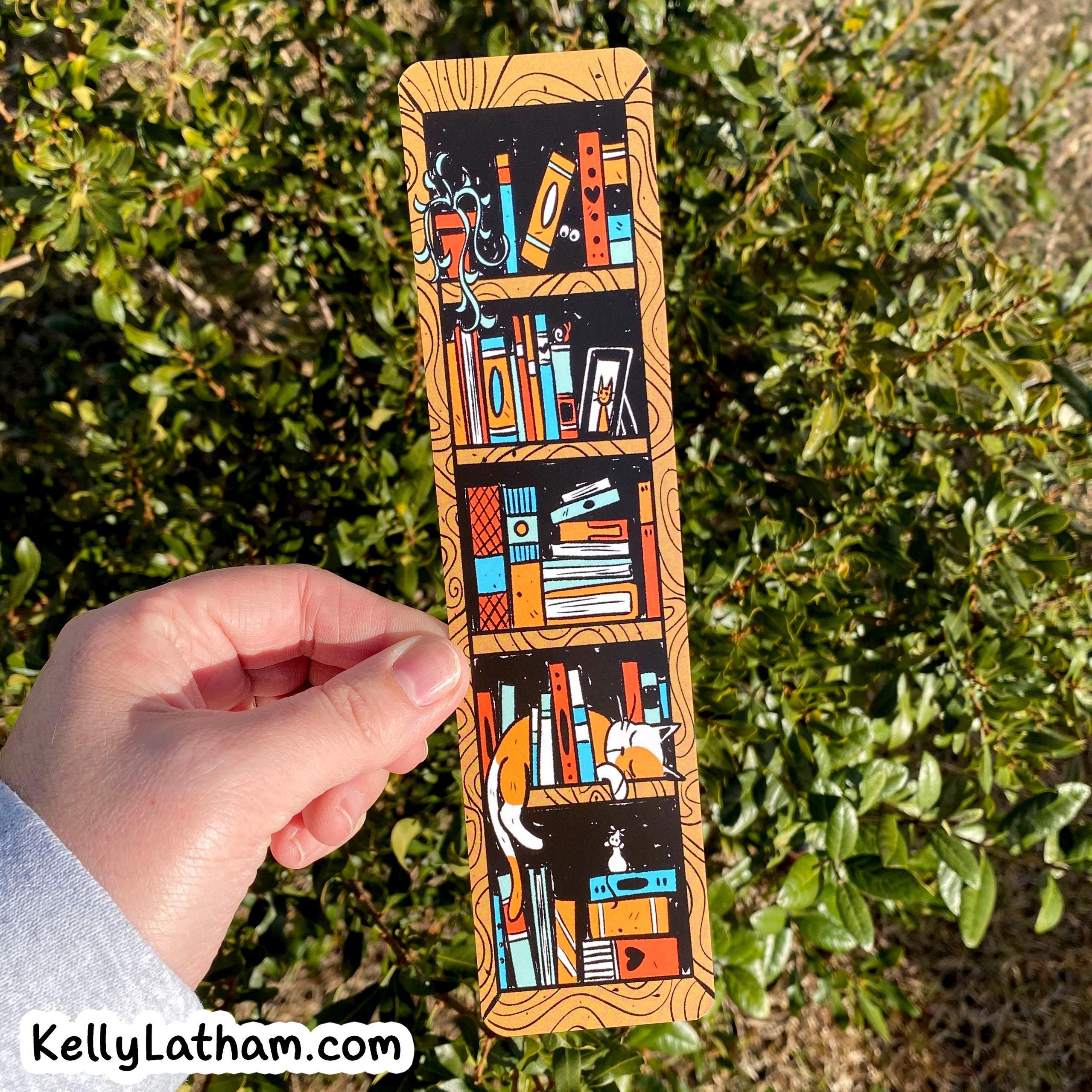 A hand holds a rectangular wooden bookplate featuring a colorful illustration of bookshelves filled with books, set against a background of green bushes outdoors.