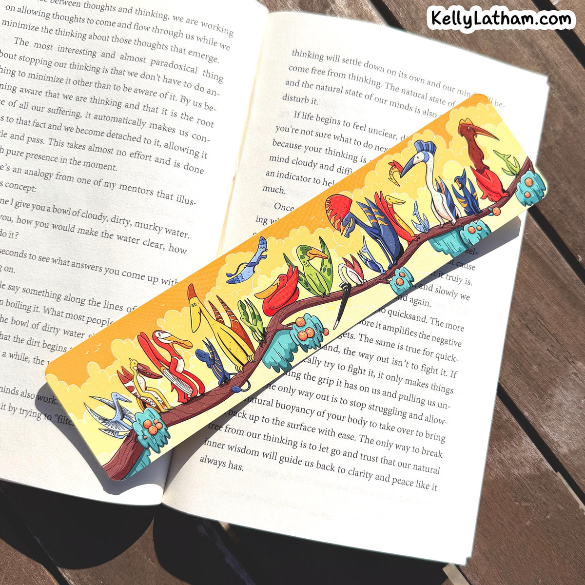 A colorful paper bookmark with a cartoon landscape scene, placed on an open book. The bookmark features a yellow background with blue sky, red sun, green hills, and blue animals.