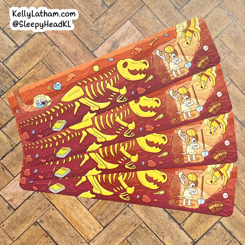 Four rectangular printed cards or sleeves laid diagonally on a wood floor, featuring a cartoon prehistoric scene with a yellow dinosaur skeleton, a paleontologist, rocks, and small book-like items. A white text watermark in the top-left reads KellyLatham.