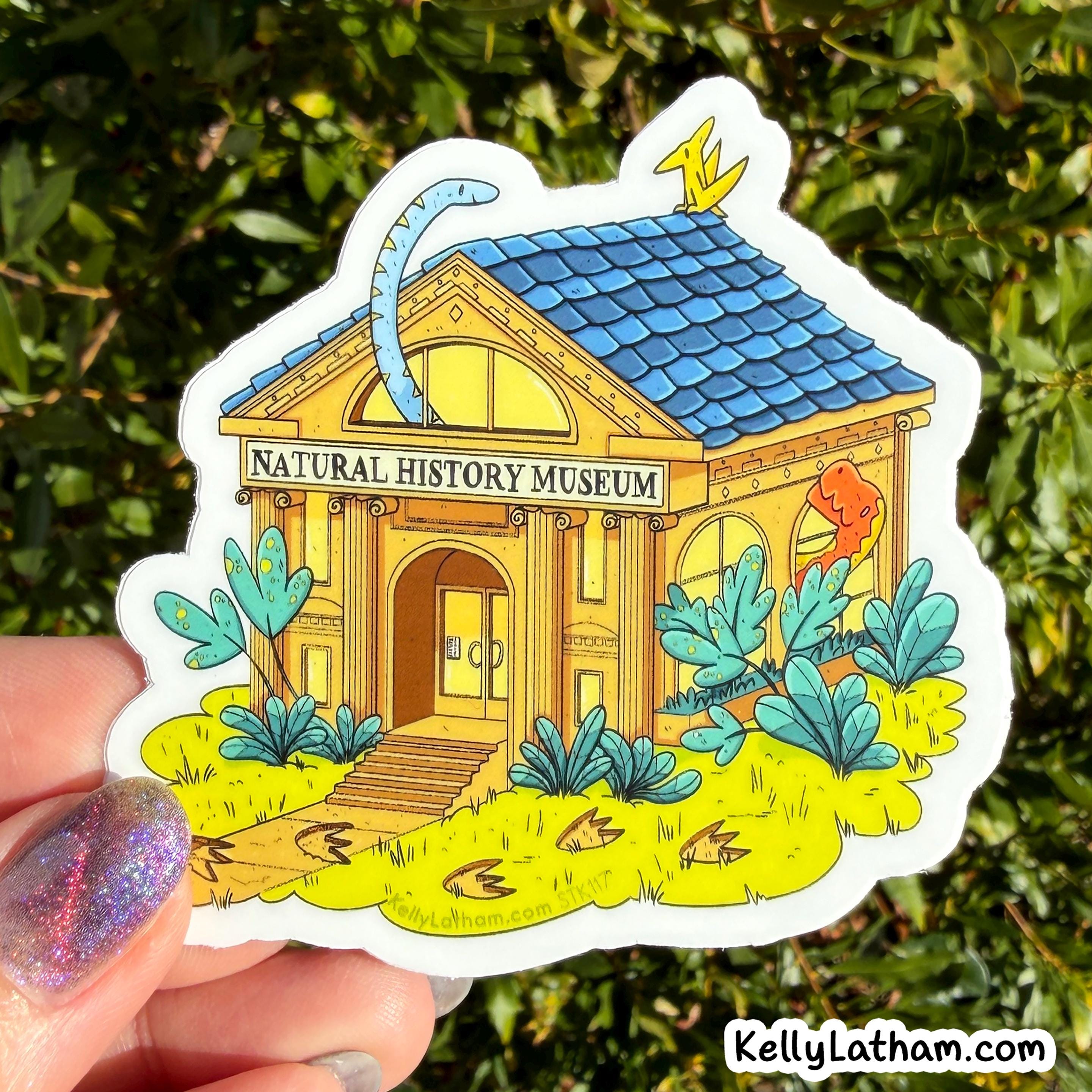 A die-cut sticker depicting a cartoon 'Natural History Museum' with a blue roof, yellow walls, and dinosaur motifs, held by a hand with glittery nail polish against a background of green foliage.