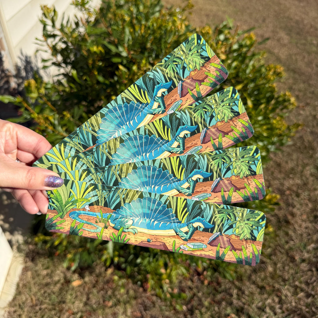 A hand holds a fanned-out bookmarks featuring a colorful tropical dinosaur print, set against a background of green bushes and dry grass.