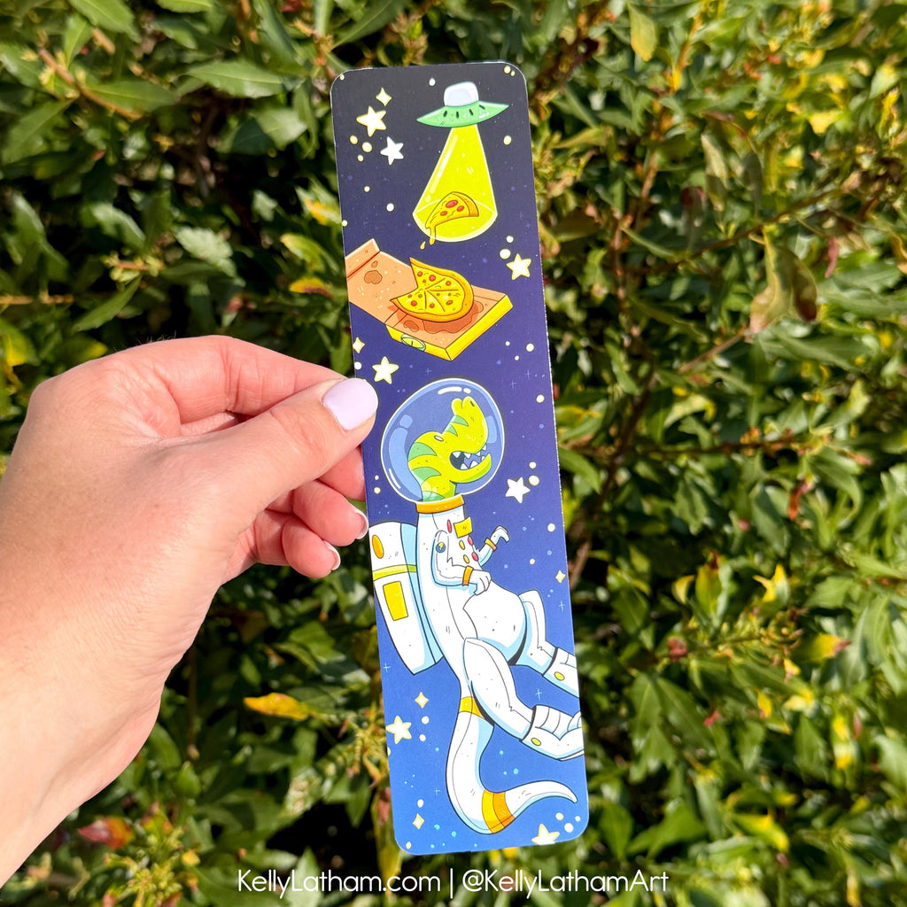 A hand holds a long rectangular printed card or bookmark featuring a whimsical space scene with a white astronaut, green dinosaur, UFO, and pizza slices on a dark blue starry background, in front of green foliage.