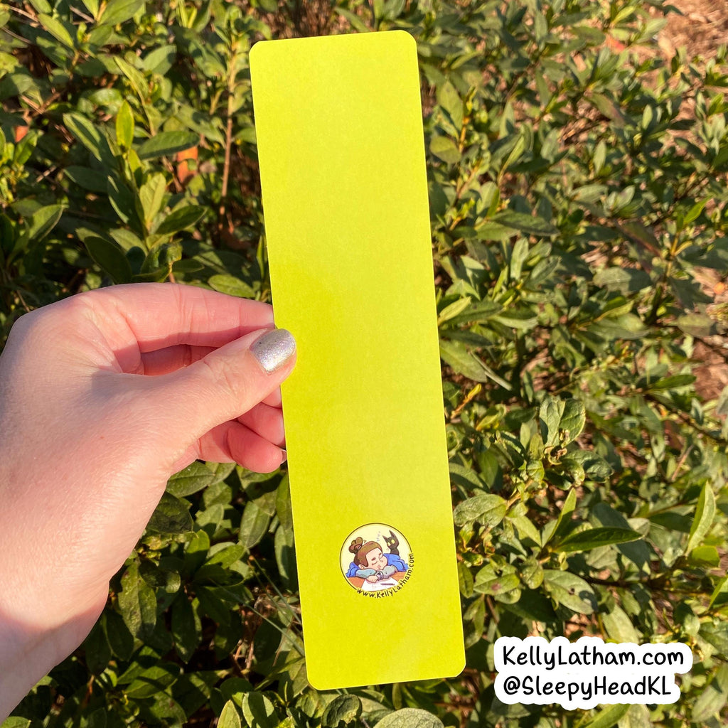 A hand holds a long, narrow neon yellow-green card or sticker outdoors in front of green bushes. The card has a small circular cartoon illustration near the bottom and a white watermark in the lower right corner reading KellyLatham.