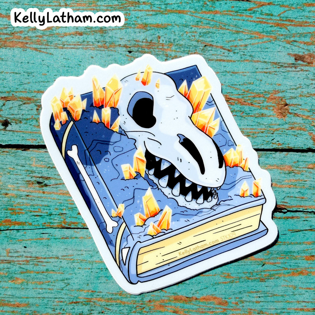 A die-cut sticker featuring a blue book with a white skull and sharp teeth, surrounded by glowing orange crystals, placed on a distressed teal wooden surface. The brand name KellyLatham.