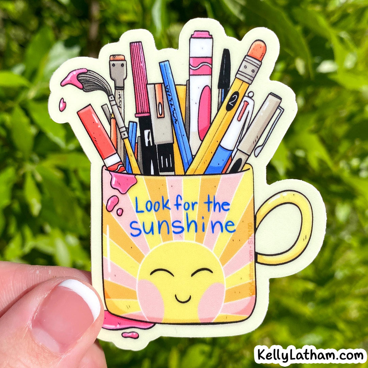 A die-cut sticker held by a hand against a blurred green foliage background. The sticker features a yellow sun face inside a pink and yellow mug with the text 'Look for the sunshine', with colorful art supplies like paintbrushes, markers, and pencils emerging from the top.