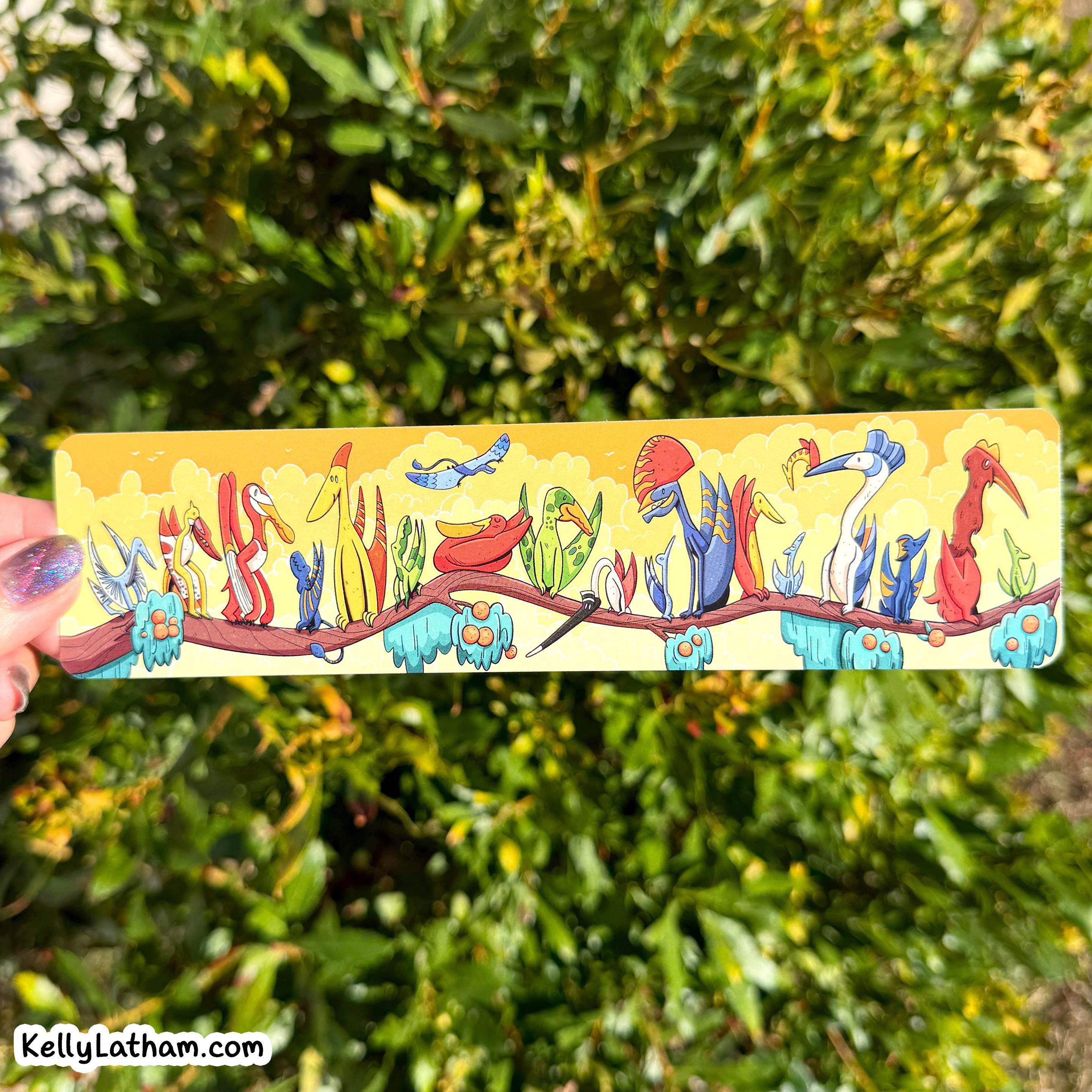A rectangular metal bar with a colorful illustration of stylized birds perched on a branch, held by a hand against a blurred green outdoor background. A watermark in the bottom-left corner reads 'KellyLatham.