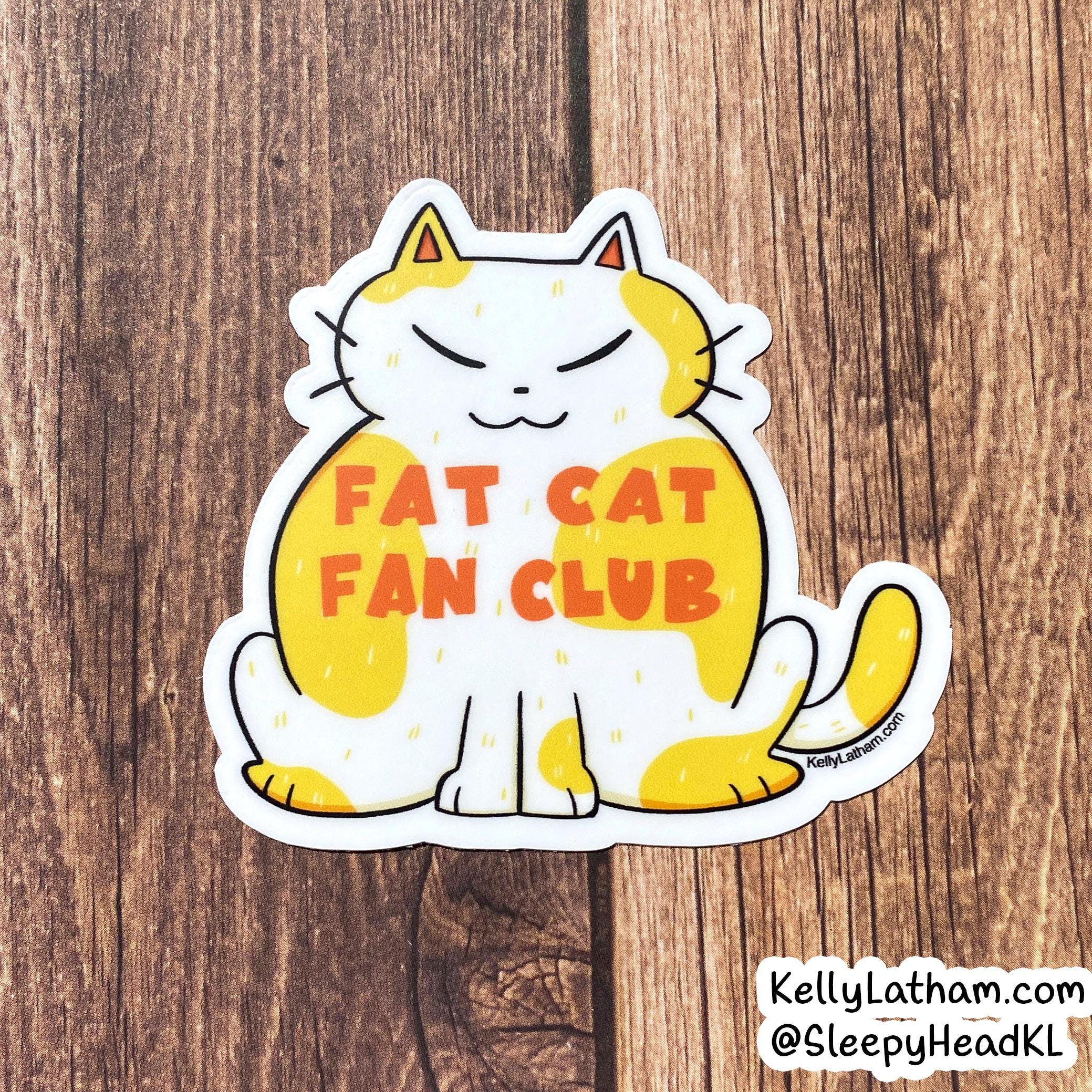 A die-cut vinyl sticker featuring a cartoon cat with yellow and white patches and the text 'FAT CAT FAN CLUB' in orange, placed on a brown wood background.