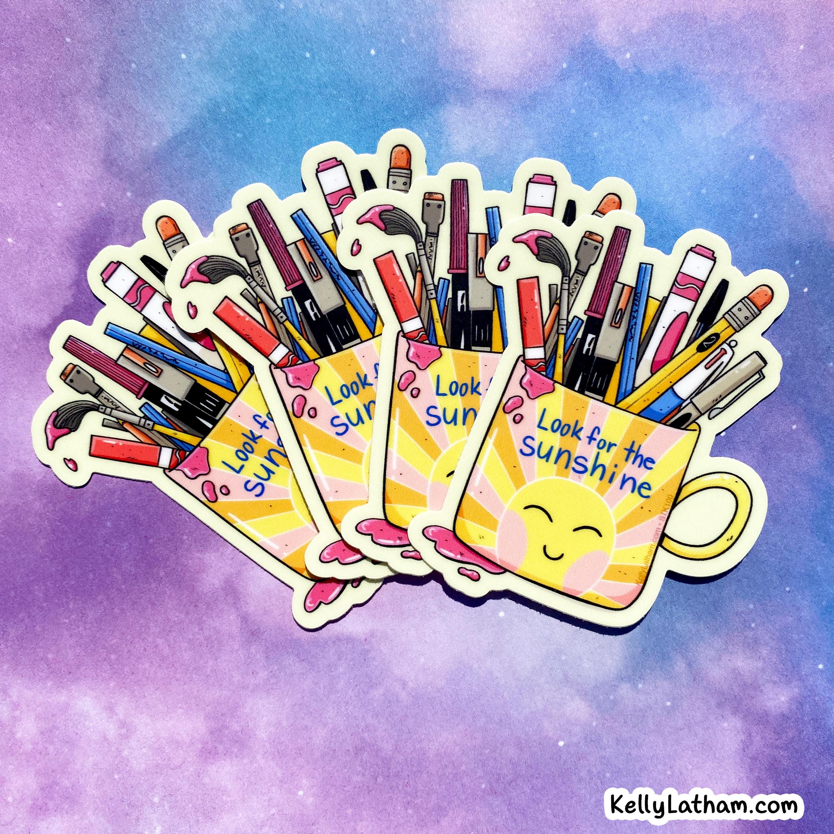 Three die-cut stickers featuring yellow smiley-face mugs filled with colorful paintbrushes and art tools, arranged in a fan shape against a purple and blue watercolor background. The stickers have the text 'Look for the Sunshine' and are shown with a small 'KellyLatham.