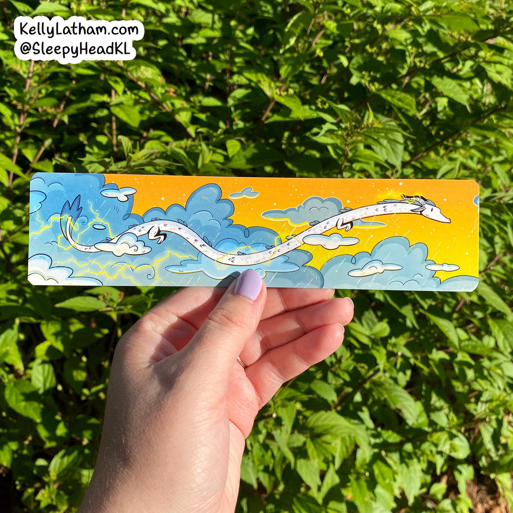 Hand holding a colorful bookmark with a dragon design against a green leafy background