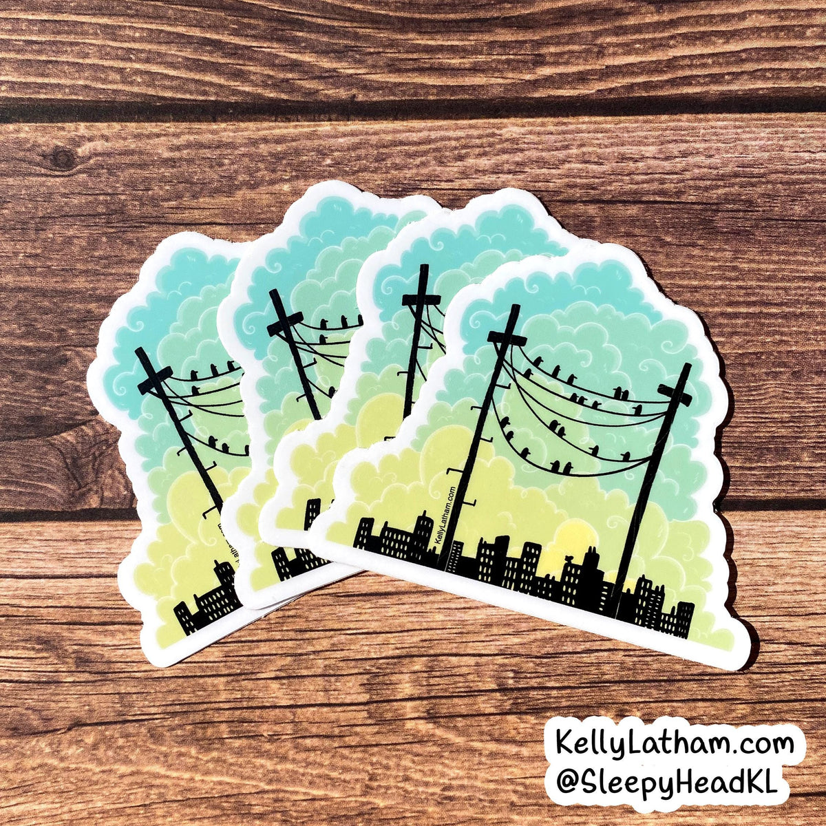 Three die-cut stickers featuring a black silhouette of a city skyline and utility poles against a yellow-to-blue gradient sky, arranged on a brown wood background. A watermark in the bottom right corner reads KellyLatham.