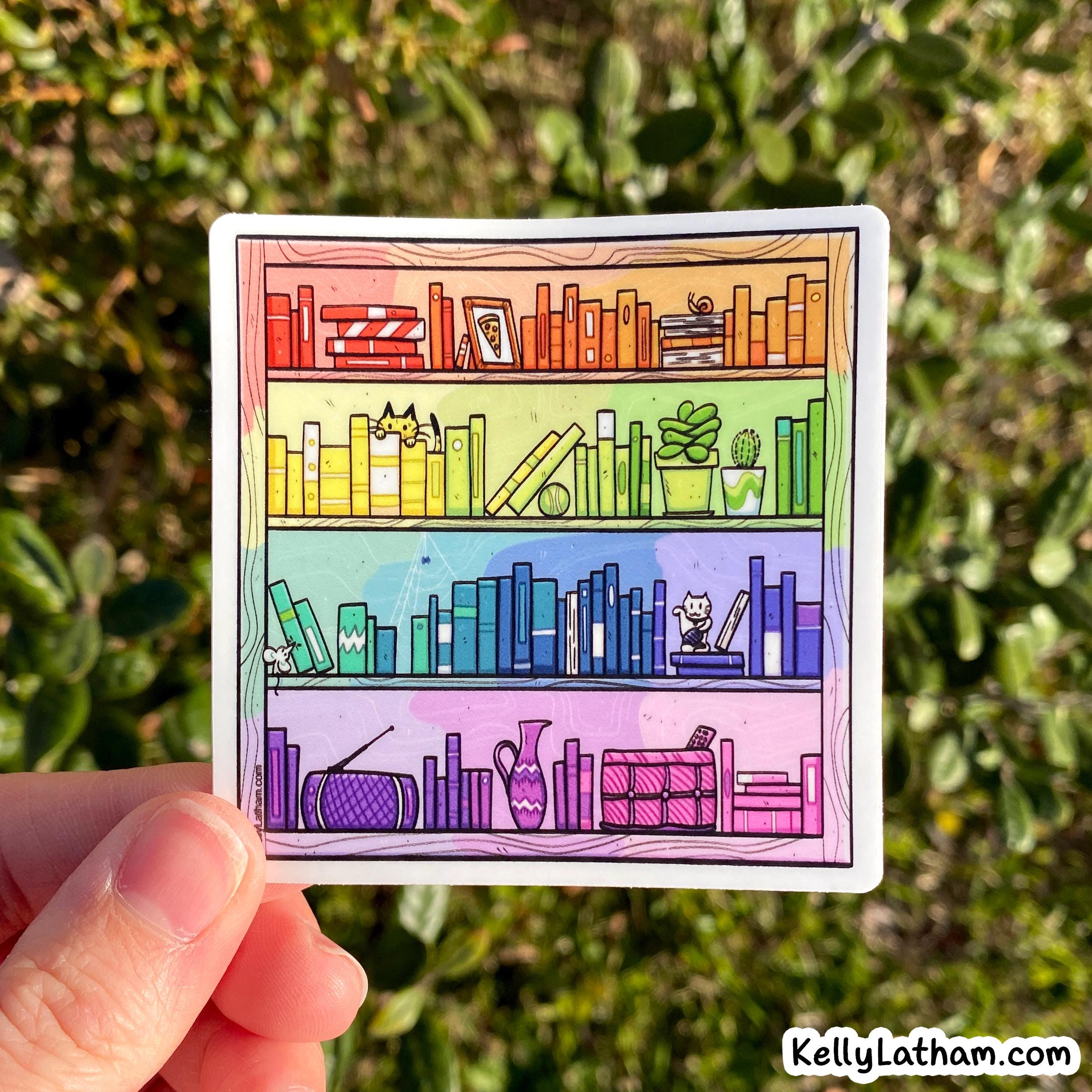 A hand holds a square sticker featuring a colorful illustrated bookshelf with books, plants, and small animals. The background is a blurred outdoor garden with green foliage.