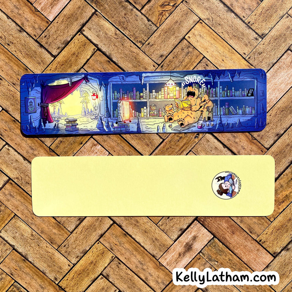 A rectangular desk mat with a detailed illustrated scene on one side and a solid pale yellow reverse side with a small circular cartoon logo, displayed on a wood herringbone floor. A 'KellyLatham.