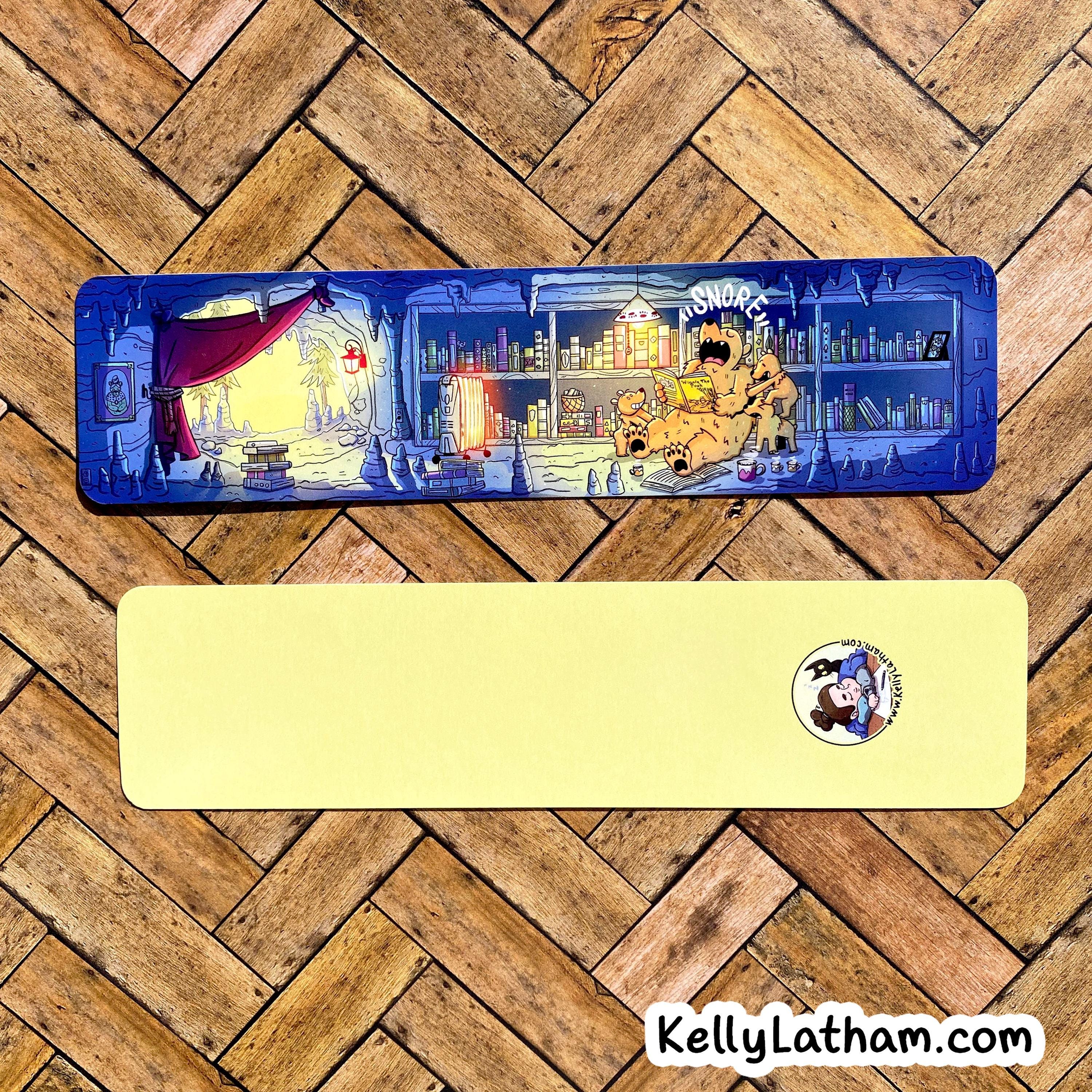 A rectangular desk mat with a detailed illustrated scene on one side and a solid pale yellow reverse side with a small circular cartoon logo, displayed on a wood herringbone floor. A 'KellyLatham.