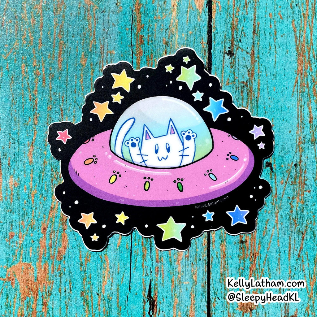 A die-cut sticker featuring a cartoon white cat in a pink UFO with a black starry space border, placed on a distressed turquoise wood background. The sticker has a small 'sleepyhead.