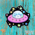 A die-cut sticker featuring a cartoon white cat in a pink UFO with a black starry space border, placed on a distressed turquoise wood background. The sticker has a small 'sleepyhead.