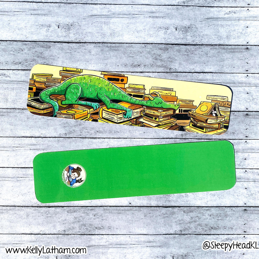 A green rectangular mouse pad with rounded corners is shown from two angles on a gray wood plank background. The top view shows a colorful illustration of a green chameleon crawling over stacks of books, with a small circular character sticker on the bottom left.