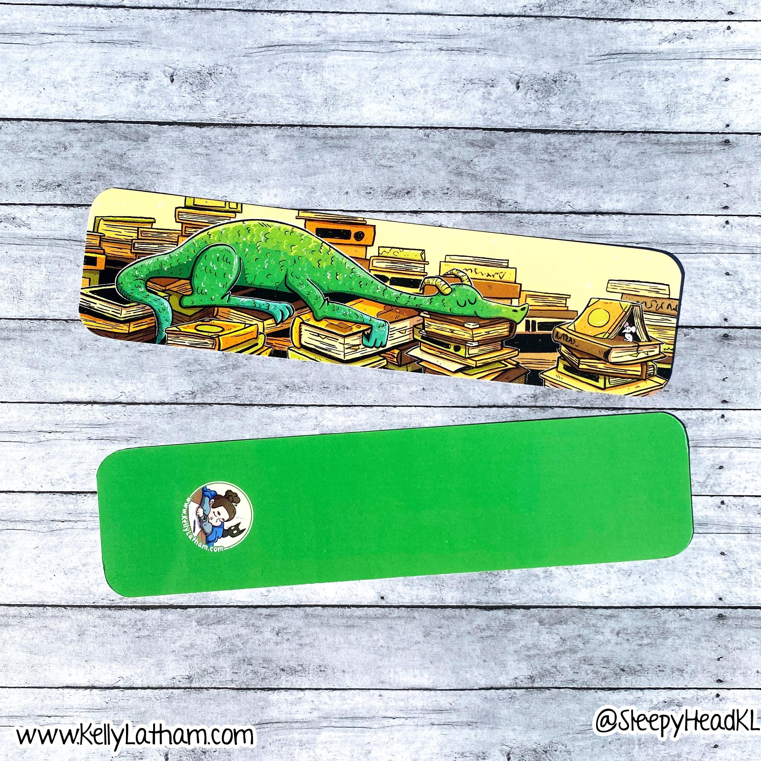 A green rectangular mouse pad with rounded corners is shown from two angles on a gray wood plank background. The top view shows a colorful illustration of a green chameleon crawling over stacks of books, with a small circular character sticker on the bottom left.