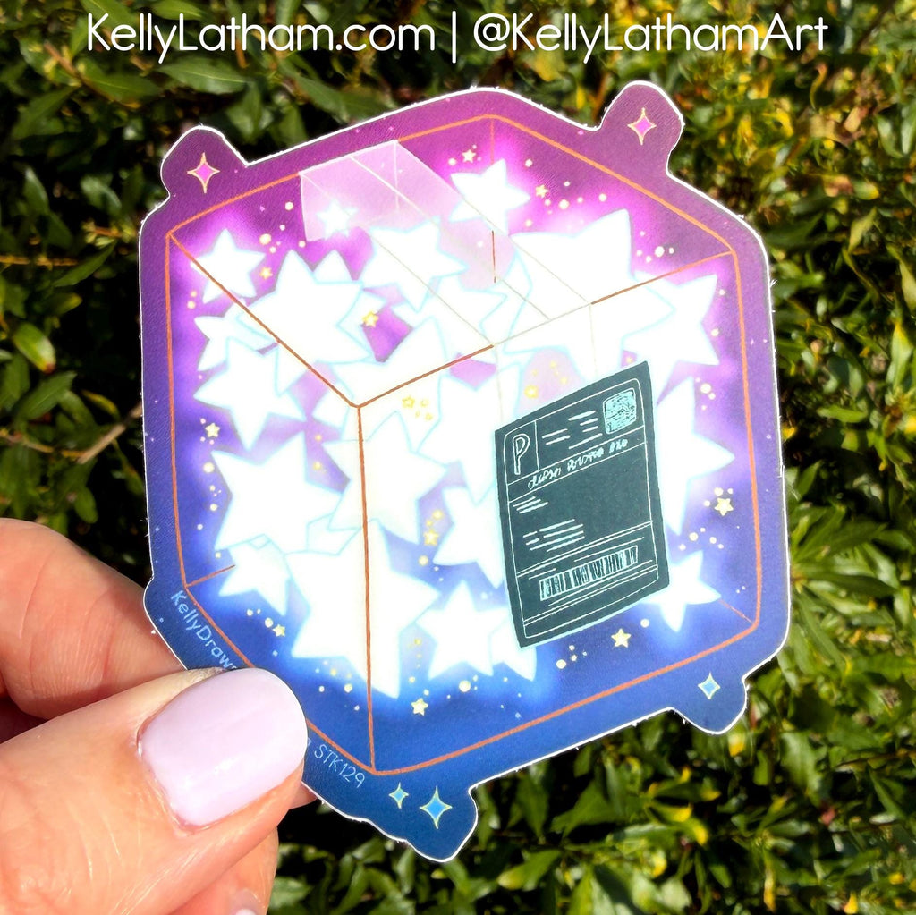 A hand with light purple nail polish holds a die-cut sticker featuring a 3D cube made of glowing white stars on a purple and blue cosmic background. The sticker has a dark green boarding pass design in the center.