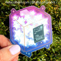 A hand with light purple nail polish holds a die-cut sticker featuring a 3D cube made of glowing white stars on a purple and blue cosmic background. The sticker has a dark green boarding pass design in the center.