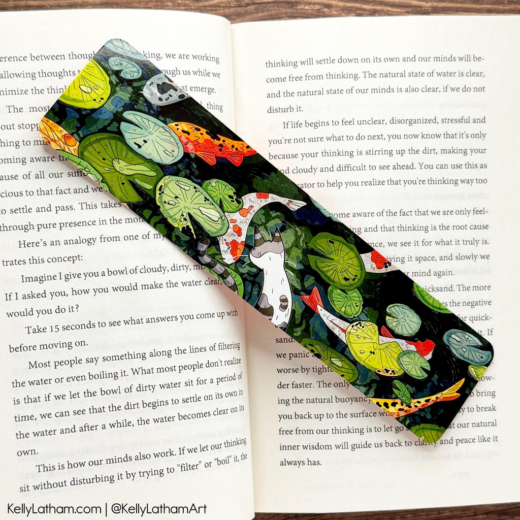 Colorful koi fish-themed bookmark with lily pads and a white and grey cat looking into the water on an open book